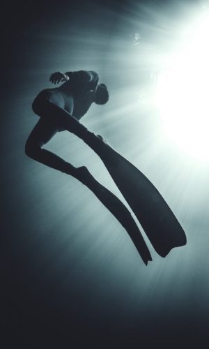 Low angle underwater view of diver wearing wet suit and flippers, sunlight filtering through from above.